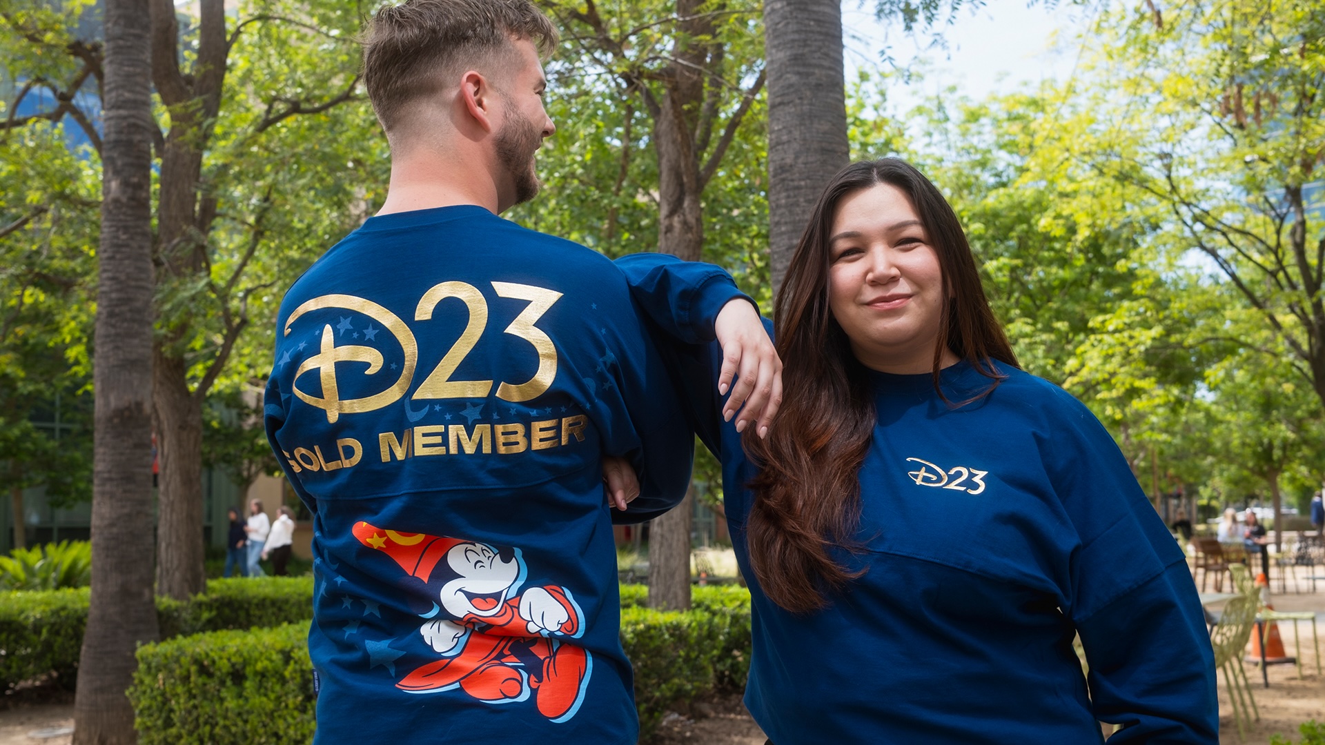 wo people wear blue D23 Gold Member Sorcerer Mickey Mouse Spirit Jerseys, featuring large gold D23 branding and Sorcerer Mickey artwork.