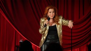 In an image from the new Hulu comedy special Lisa Ann Walter: It Was an Accident, actress and comedian Walter stands on stage, one hand holding a mic and the other arm resting on the mic stand. She&rsquo;s wearing a gold lam&eacute; blazer.