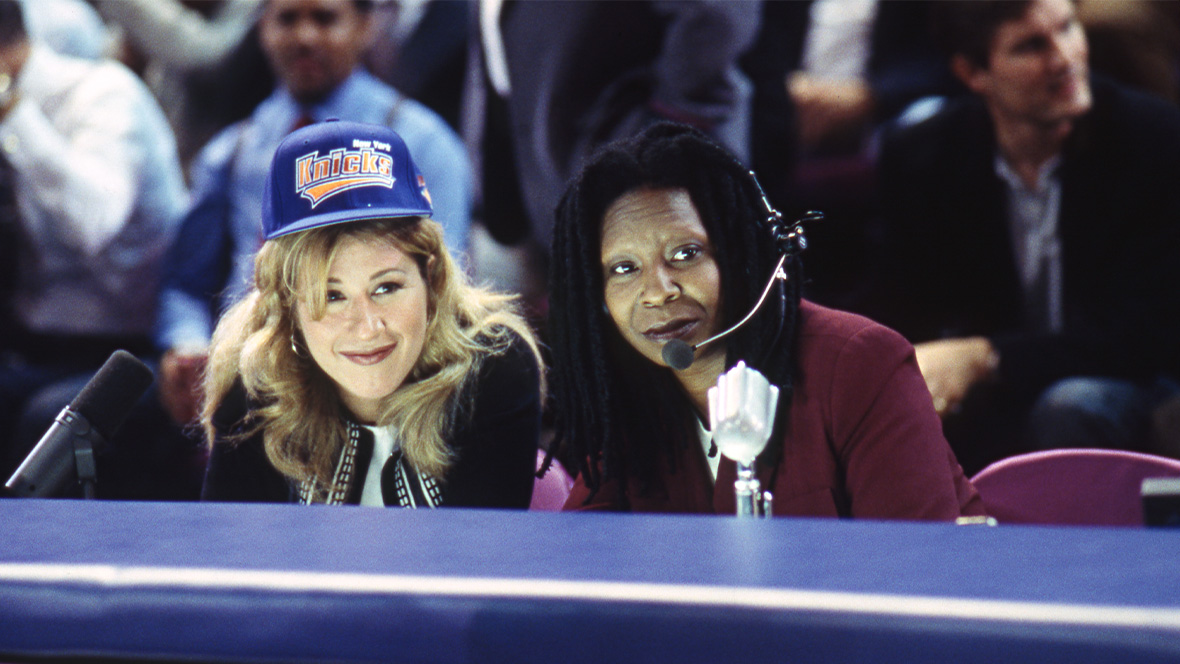 In an image from 1996&rsquo;s Eddie, Claudine (Lisa Ann Walter) and Eddie (Disney Legend Whoopi Goldberg) sit side-by-side behind a table during a New York Knicks game. Eddie is wearing a headset with a microphone.