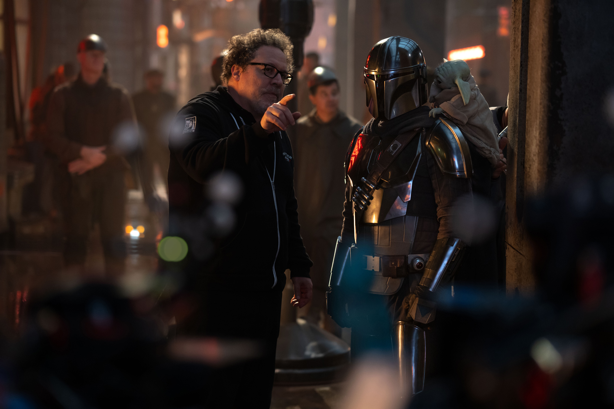 An image of Disney Legend Jon Favreau on the set of Star Wars: The Mandalorian and Grogu. To his right is Pedro Pascal in costume as Din Djarin, with Grogu on his back; other actors and crew members can be seen in the background.