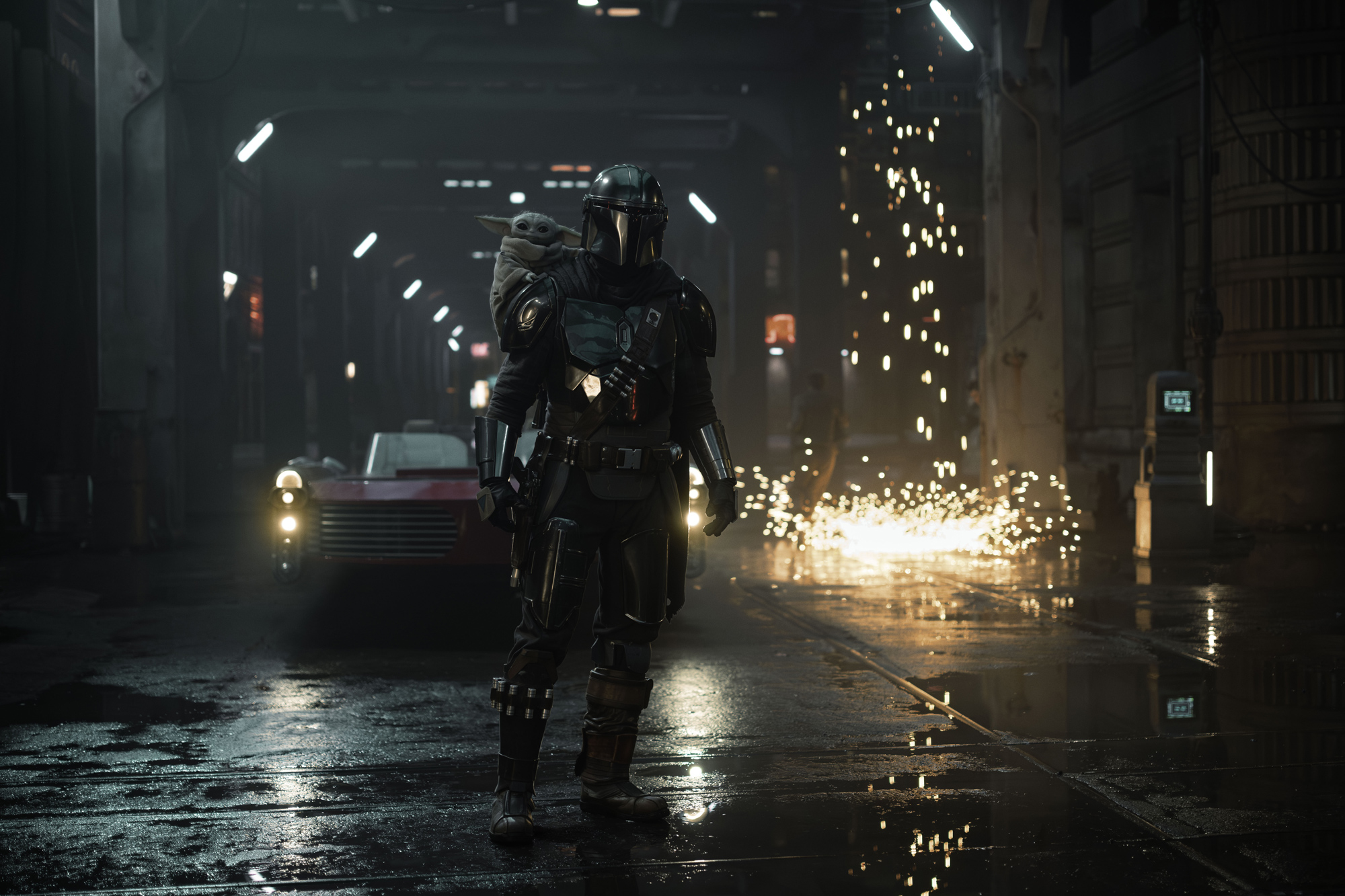 An image from Lucasfilm&rsquo;s Star Wars: The Mandalorian and Grogu, featuring Pedro Pascal as Din Djarin, with Grogu being carried on his back. They are walking down a street; a vehicle can be seen behind them, as well as a waterfall of sparks to the right of the vehicle.