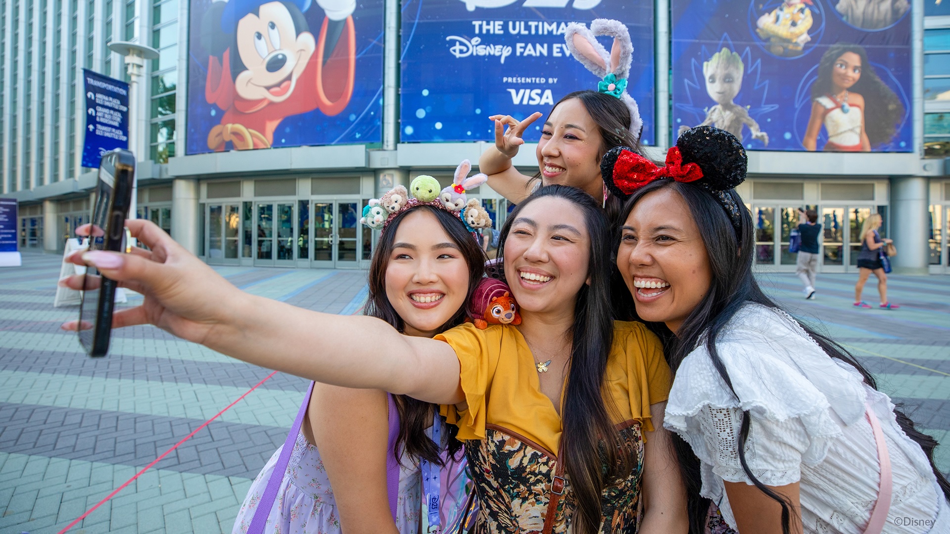 Fans wearing Disney-themed accessories take a selfie outside the Anaheim Convention Center under a large banner for D23: The Ultimate Disney Fan Event.