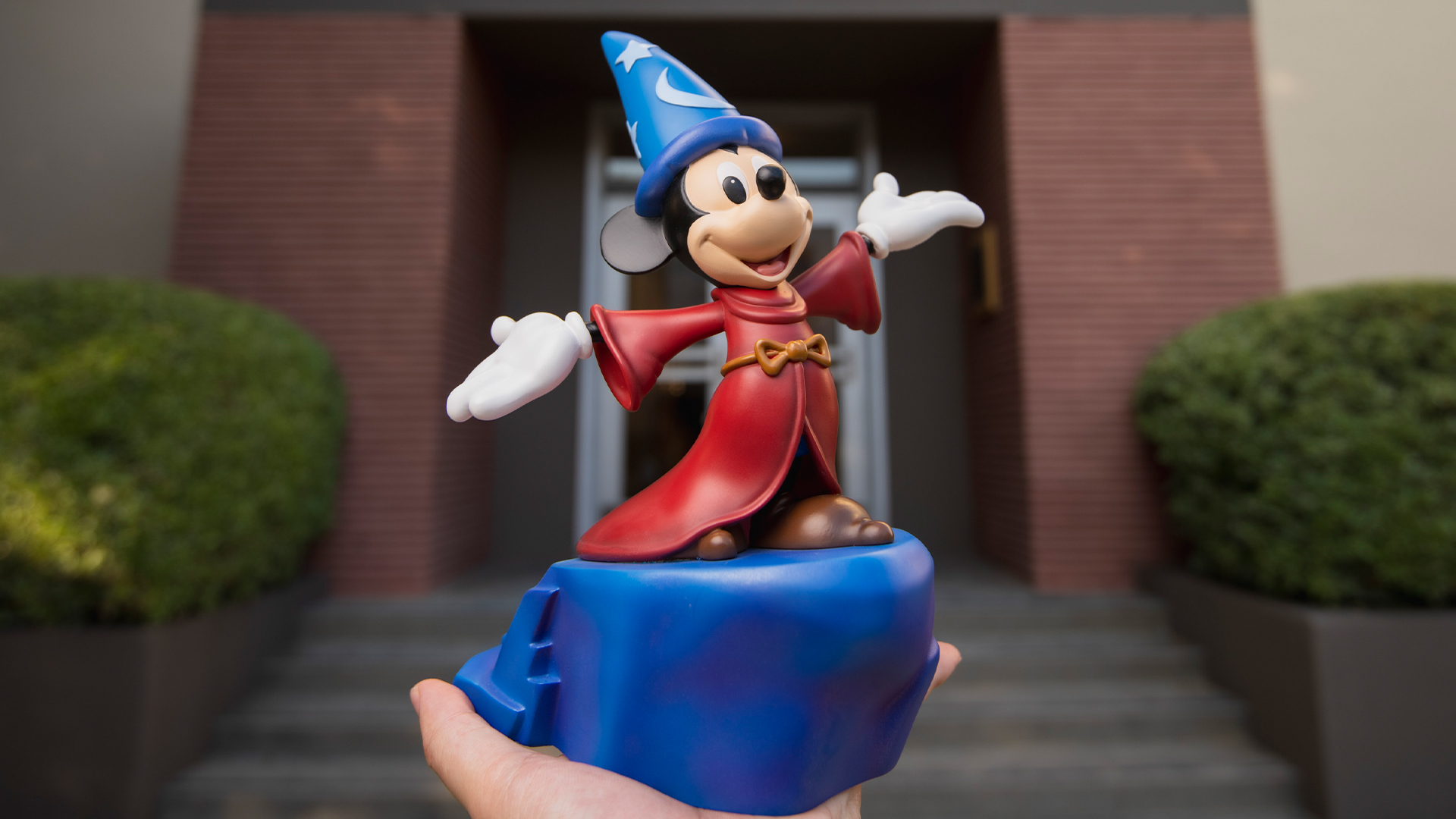A Sorcerer Mickey Mouse figurine in a red robe and blue hat is held up in front of a building labeled ANIMATION.