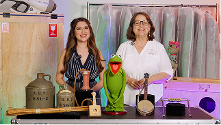 Two people stand behind Muppet character Kermit the Frog and various props in a backstage setting with costume bags and storage crates.