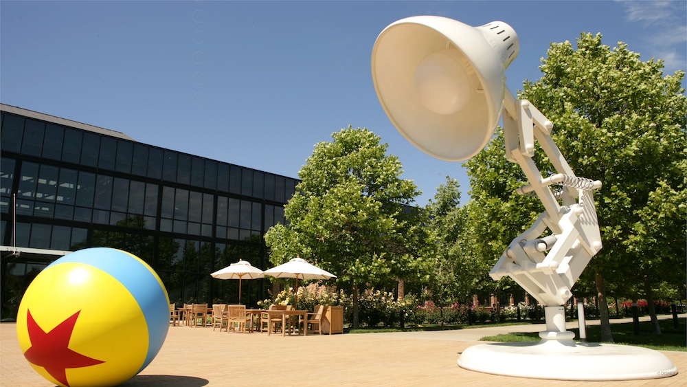 The giant Pixar Lamp and Luxo Ball stand outdoors in front of a glass building with patio tables and chairs.