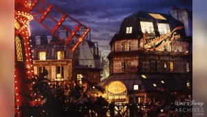 A nighttime view of a romantic Parisian street scene, featuring the red-lit windmill of the Moulin Rouge on the left and old-style buildings with glowing windows. One building has a neon sign that reads &ldquo;L&rsquo;amour fou Lingerie&rdquo; and another displays &ldquo;Hotel Blanche.&rdquo; The sky is dark blue with clouds, and the overall ambiance is dreamy and nostalgic.