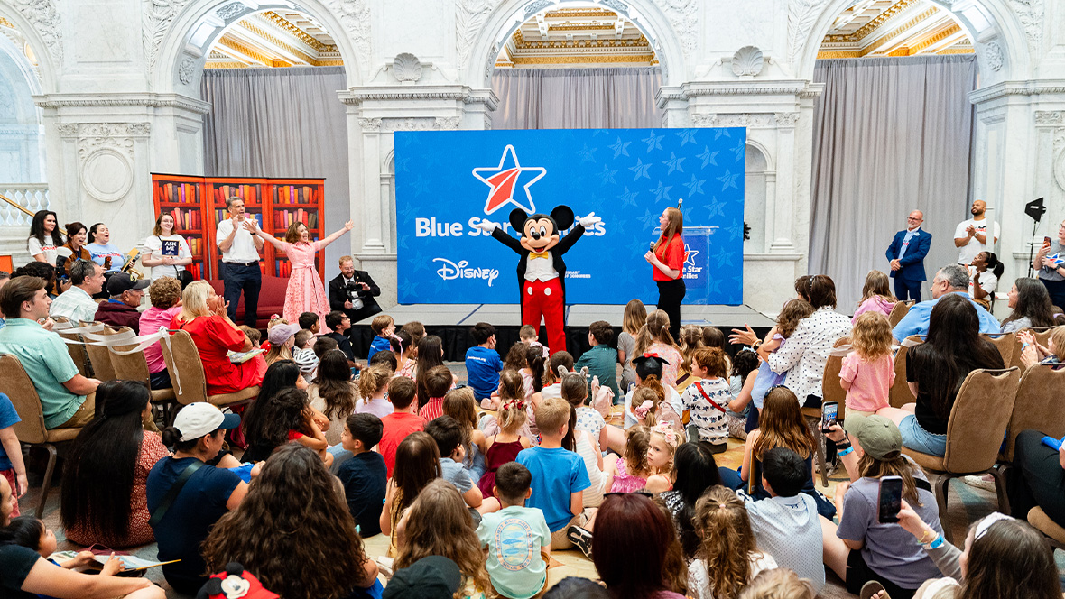 An image of Mickey Mouse celebrating with Blue Star Families—including children sitting in a semi-circle—at an event.