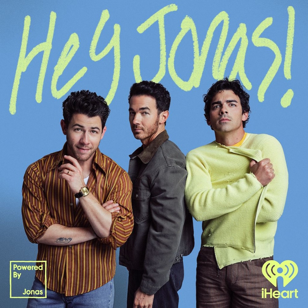 The Jonas Brothers stand together posing in front of a blue background with "Hey Jonas!" written above and iHeart logo visible.