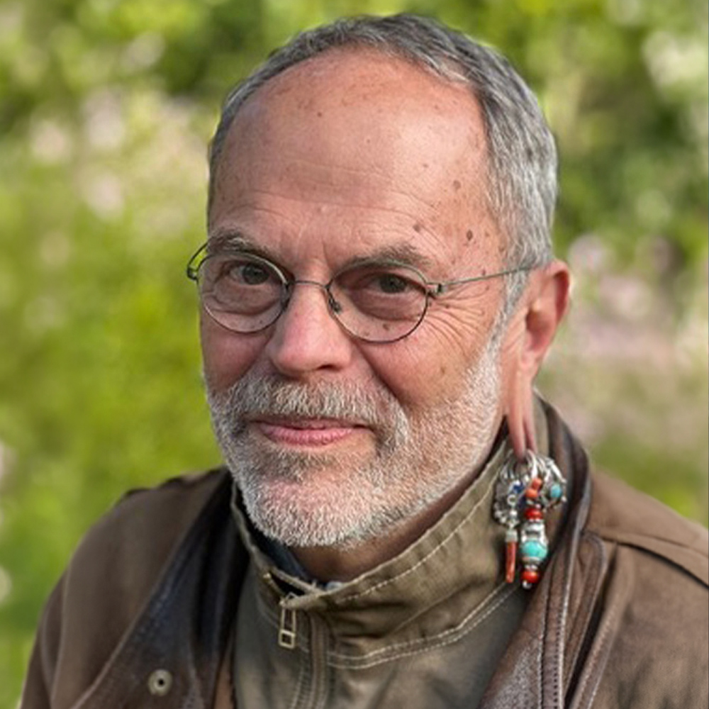 Portrait of a person wearing glasses, a brown jacket, and decorative earrings outdoors against a softly blurred green background.