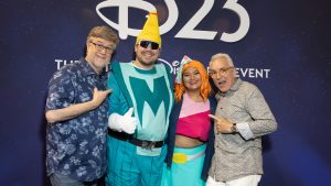 Four D23 Expo attendees posing in front of a &ldquo;D23&rdquo; backdrop, including two colorful Disney cosplay characters.