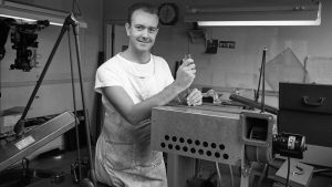 Don Iwerks adjusts a mechanical device at a workbench inside a studio filled with cameras, tools, and equipment.