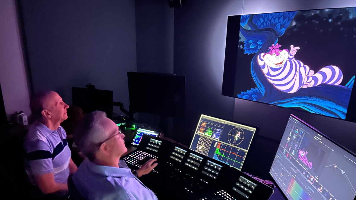 Two people use color-grading workstations while adjusting a Cheshire Cat scene from Disney&rsquo;s Alice in Wonderland on studio monitors.