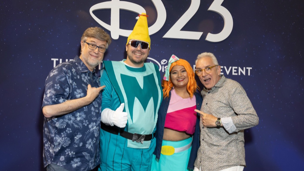Four D23 Expo attendees posing in front of a &ldquo;D23&rdquo; backdrop, including two colorful Disney cosplay characters.