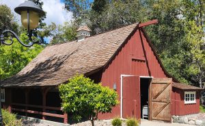 Red barn-style building with an open wooden door, brown shingled roof, and white-trimmed windows. The structure is surrounded by trees, bushes, and a landscaped pathway in daylight.
