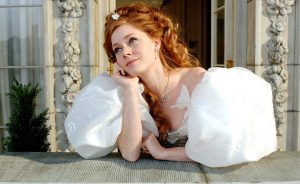 Giselle from Enchanted leans on a stone balcony in front of an elegant building with carved floral stonework and tall windows. The character wears a shimmering white ball gown with oversized puff sleeves, jewelry accents, and a butterfly embellishment near the neckline. Warm daylight illuminates the scene.
