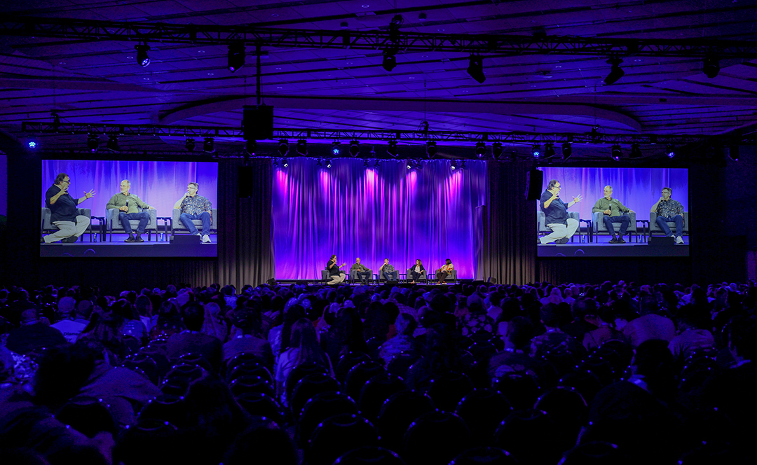 Audience members watch a panel discussion on a large stage illuminated with purple lighting, with the speakers shown on two large screens.