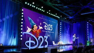Speaker presents on stage beside large D23 backdrop reading &ldquo;August 14&ndash;16, 2026, Anaheim, CA&rdquo; at Disney fan event.