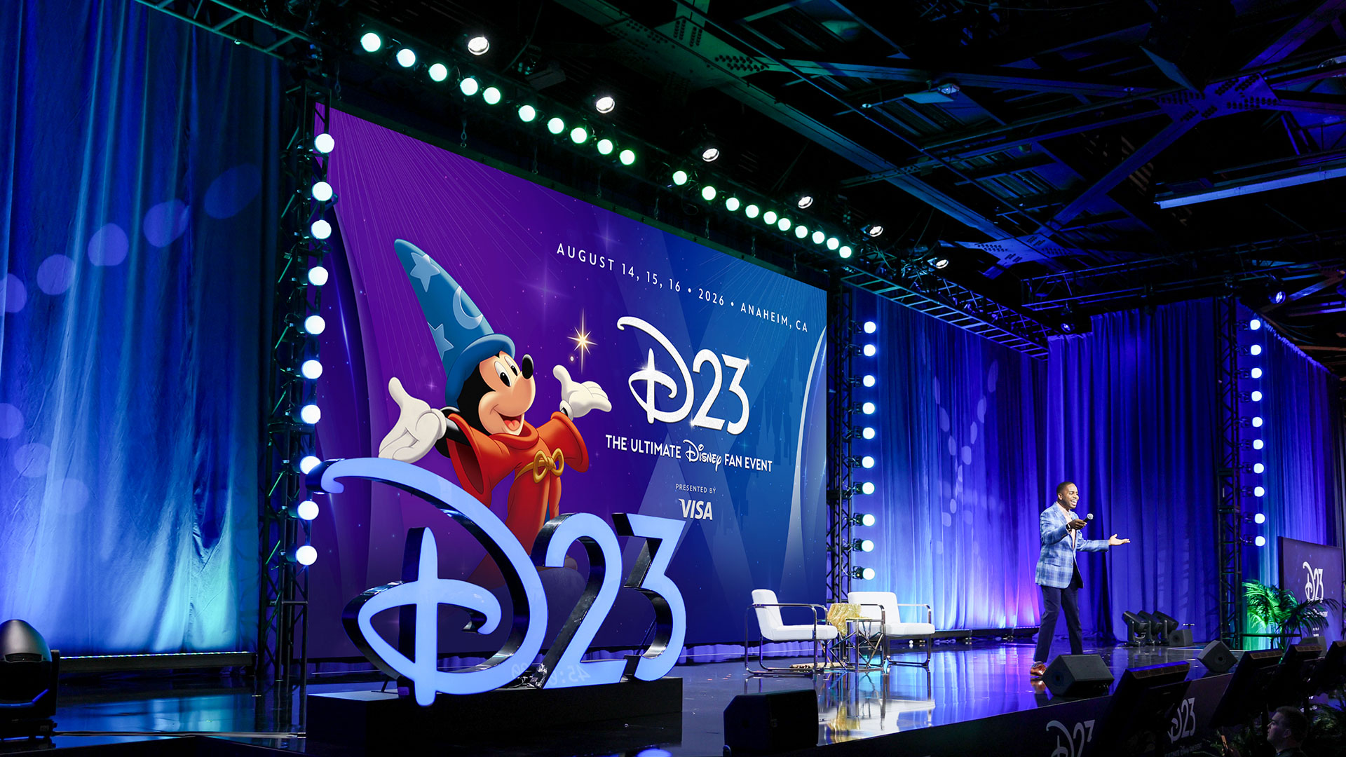 Speaker presents on stage beside large D23 backdrop reading &ldquo;August 14&ndash;16, 2026, Anaheim, CA&rdquo; at Disney fan event.