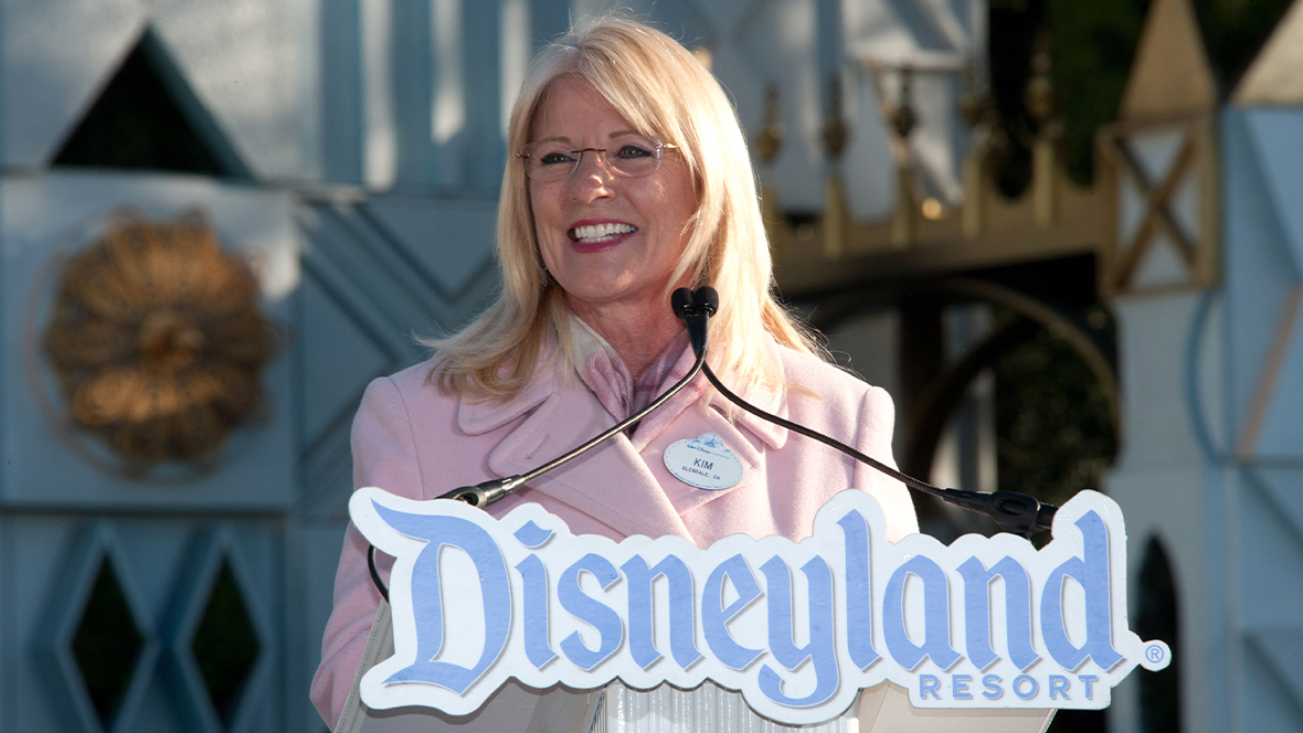 Disney Legend Kim Irvine speaks at the podium in front of "it's a small world" at Disneyland Park.