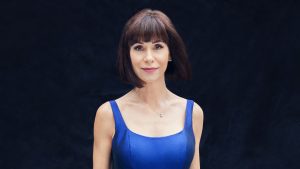 Disney Legend Susan Egan wears a violet dress and stands against a black backdrop.