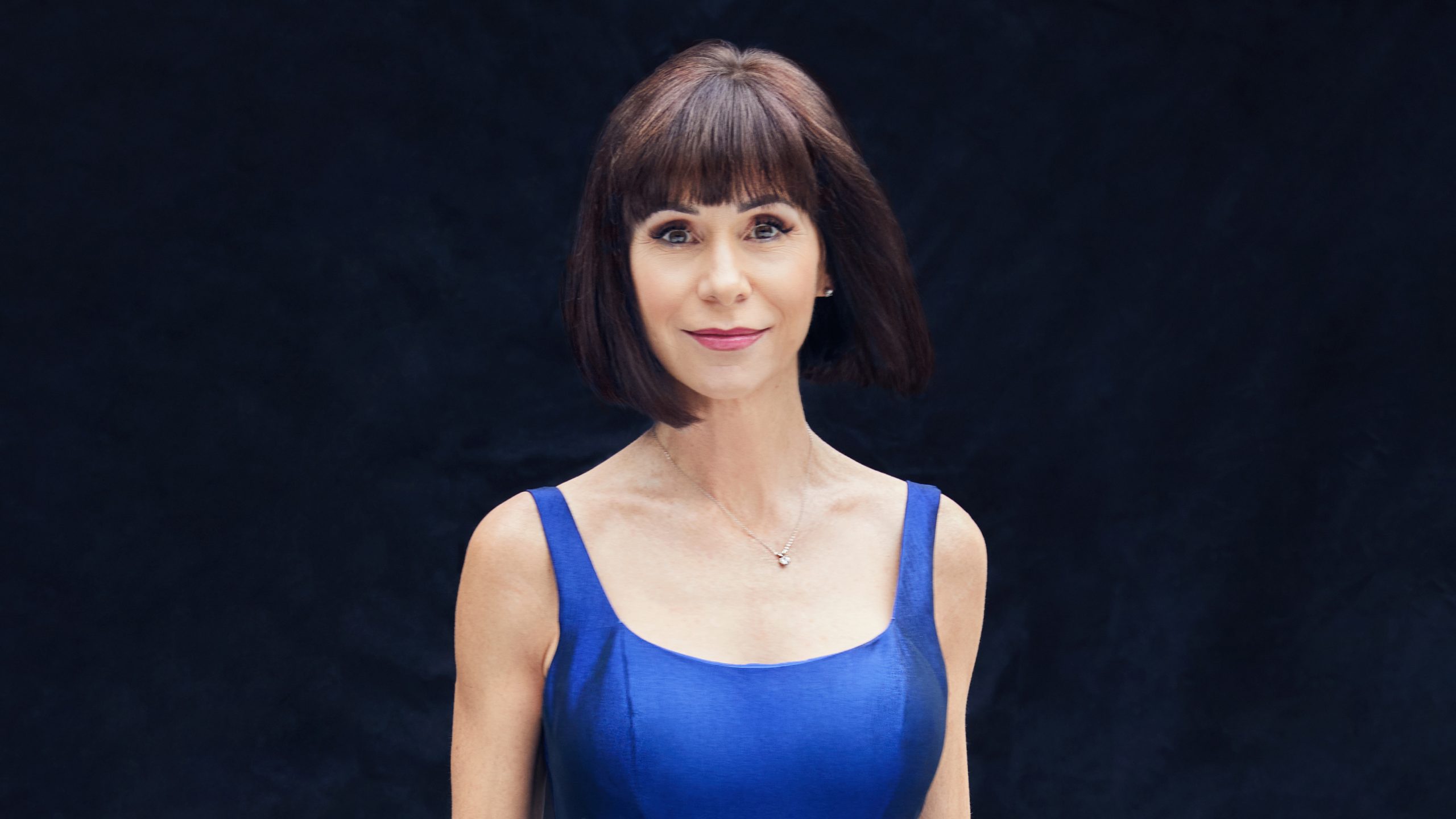 Disney Legend Susan Egan wears a violet dress and stands against a black backdrop.