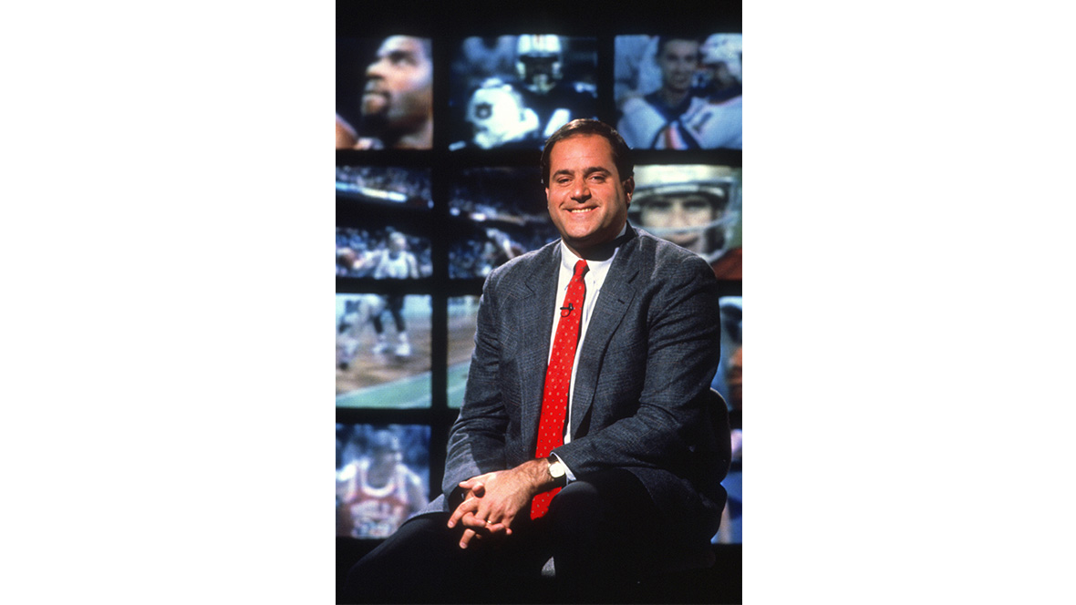 Disney Legend Chris Berman sits in front a wall of television monitors.