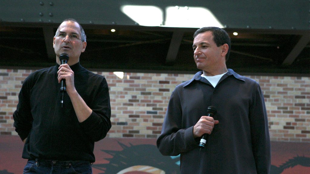 Steve Jobs and Bob Iger hold microphones while speaking to an audience during a corporate presentation event.