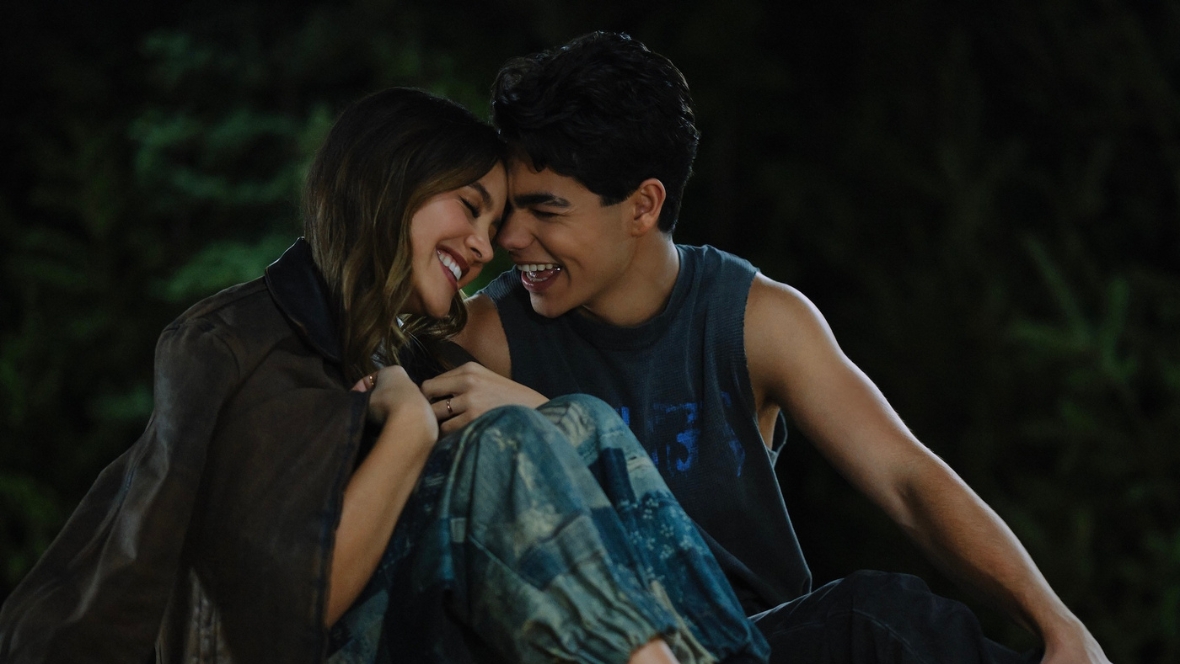 Sage, played by Liamani Segura, and Fletch, played by Malachi Barton, are laughing and touching foreheads while sitting on a dock in a scene from Camp Rock 3.