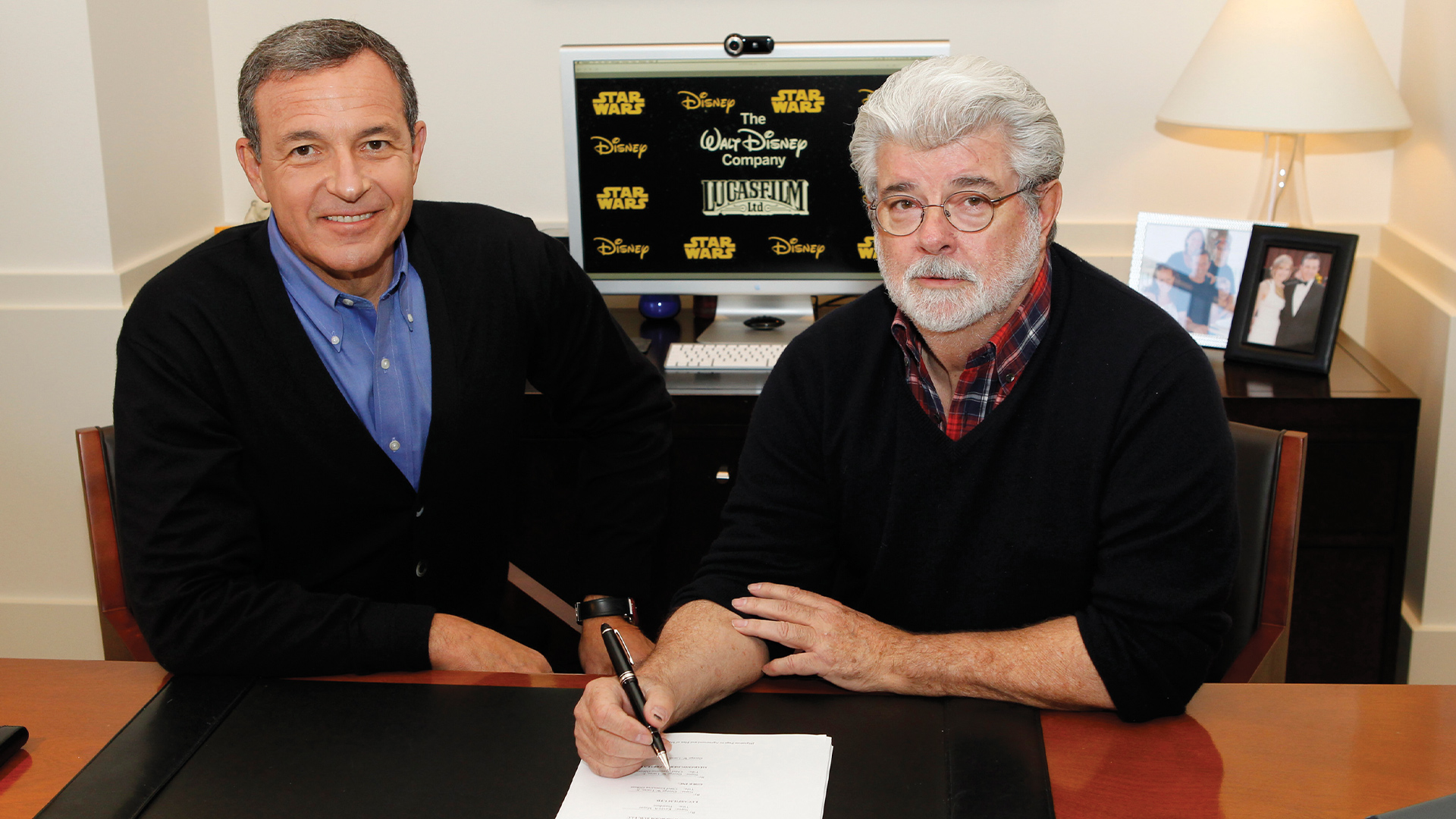 Bob Iger and George Lucas sit at a conference table as one signs a document during a corporate acquisition announcement event.