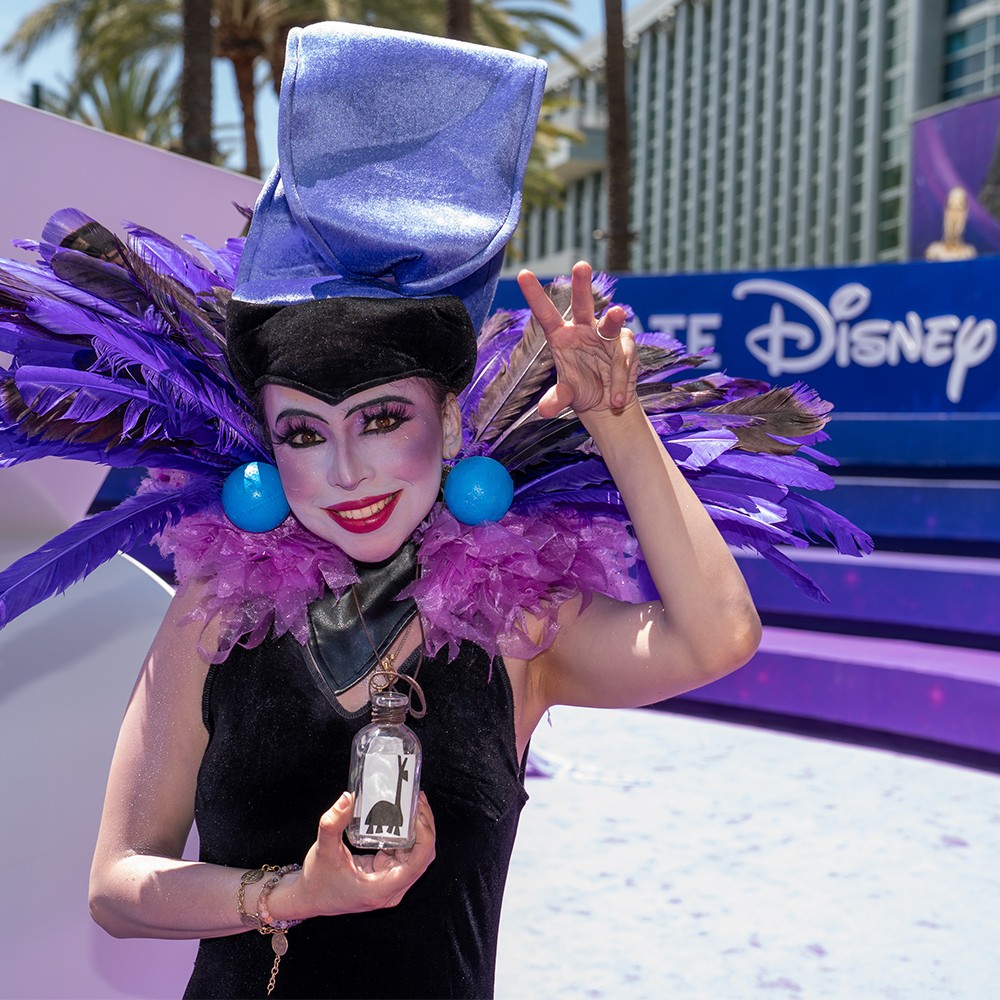 A D23 attendee cosplaying as Yzma from Disney&rsquo;s The Emperor&rsquo;s New Groove poses in front of D23: The Ultimate Disney Fan Event signage.