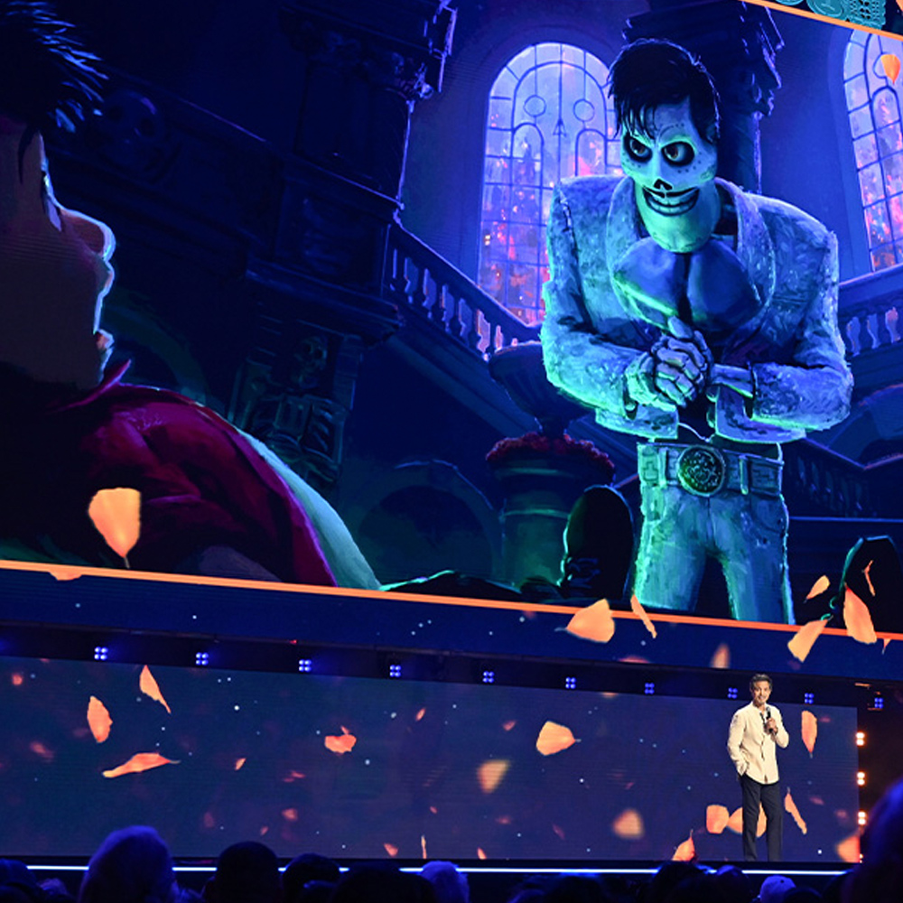 An image of Benjamin Bratt on stage, with an image of Coco 2&rsquo;s Ernesto de la Cruz in the background, at the Honda Center during the Disney Entertainment Showcase at D23: The Ultimate Disney Fan Event 2026.
