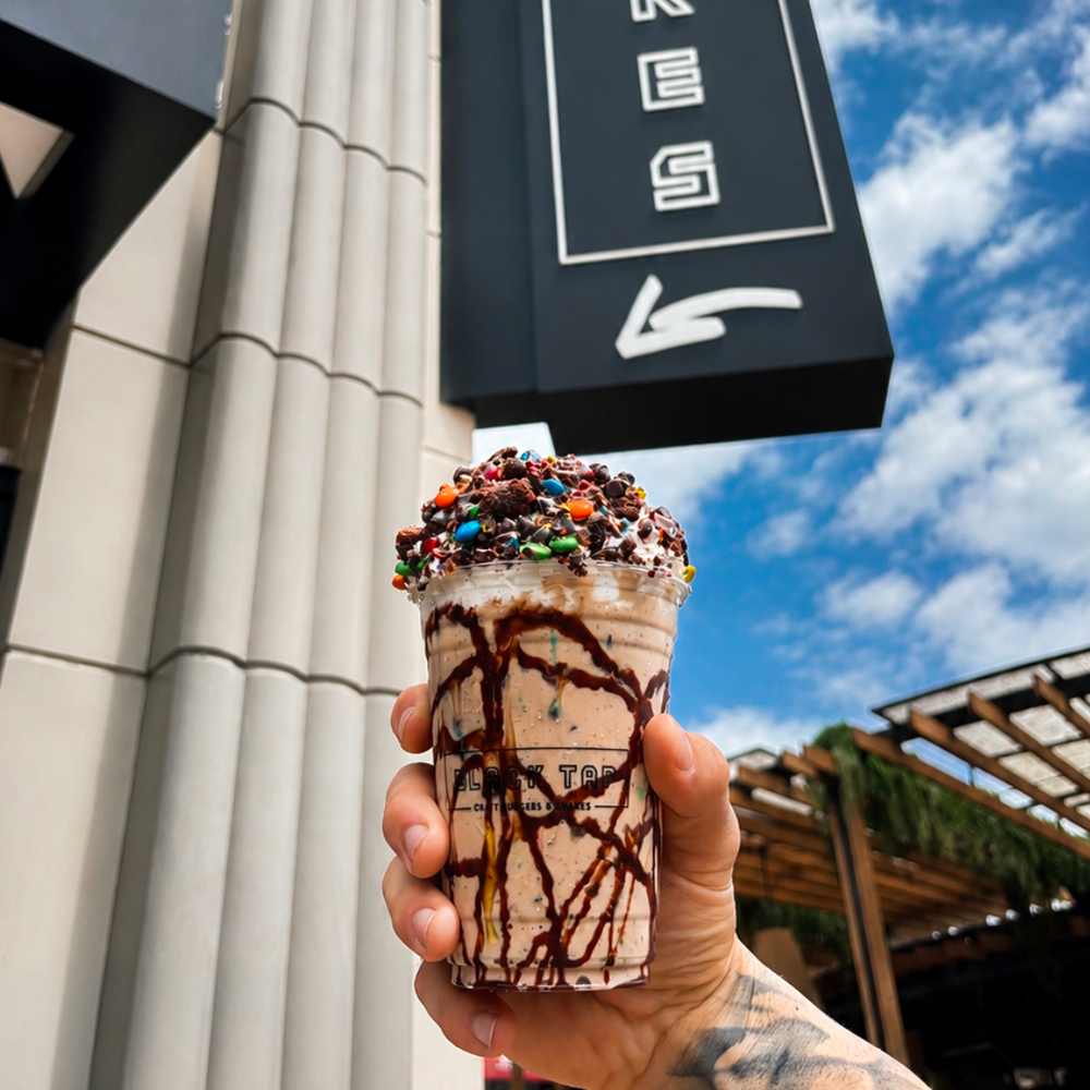 Black Tap milkshake topped with whipped cream, chocolate drizzle, candy-coated chocolates, and cookie pieces is held outside the restaurant.
