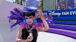 A D23 attendee cosplaying as Yzma from Disney&rsquo;s The Emperor&rsquo;s New Groove poses in front of D23: The Ultimate Disney Fan Event signage.