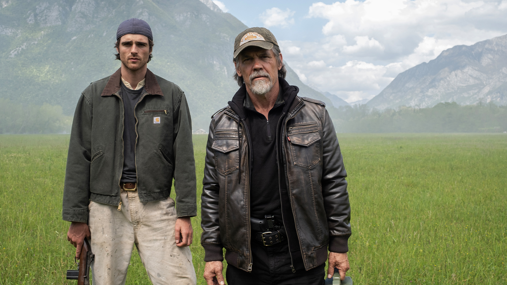 Hig, played by Jacob Elordi, and Bangley, played by Josh Brolin, stand side-by-side in a grassy field in a scene from 20th Century Studios' The Dog Stars. Hig is holding a shotgun in his right hand, while Bangley is carrying a green can of gasoline in his left hand.