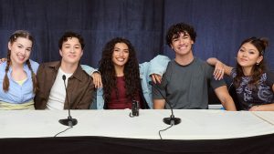 An image of the lead cast from Disney Channel&rsquo;s Hidden Heroes: A Descendants Story, including Dior Goodjohn, Momona Tamada, Jacob Rodriguez, Eva Fossaceca Smedley, and Harry Jones. They are sitting behind a table and have their arms around each other&rsquo;s shoulders.