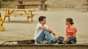 Fletch, played by Malachi Barton, and Sage, played by Liamani Segura, laugh and talk on a wooden dock in a scene from Camp Rock 3. An acoustic guitar is by Fletch's side.