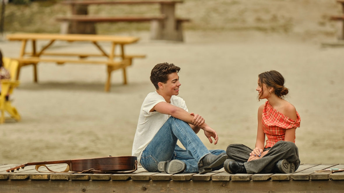 Fletch, played by Malachi Barton, and Sage, played by Liamani Segura, laugh and talk on a wooden dock in a scene from Camp Rock 3. An acoustic guitar is by Fletch's side.