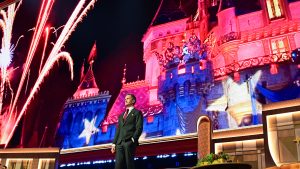 Neil Patrick Harris stands onstage during D23: The Ultimate Disney Fan Event with Disneyland fireworks and Sleeping Beauty Castle imagery displayed behind them.