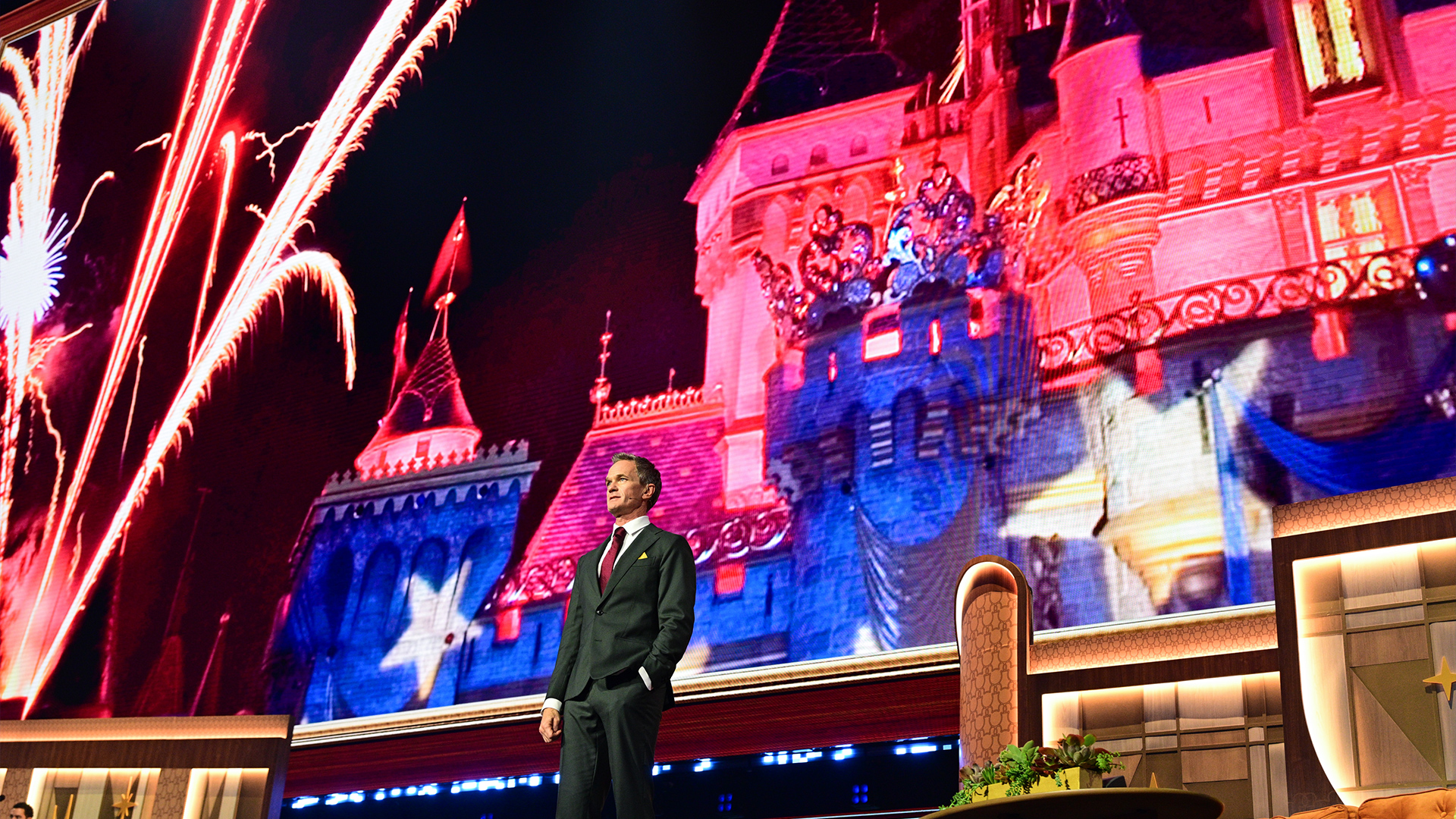 Neil Patrick Harris stands onstage during D23: The Ultimate Disney Fan Event with Disneyland fireworks and Sleeping Beauty Castle imagery displayed behind them.