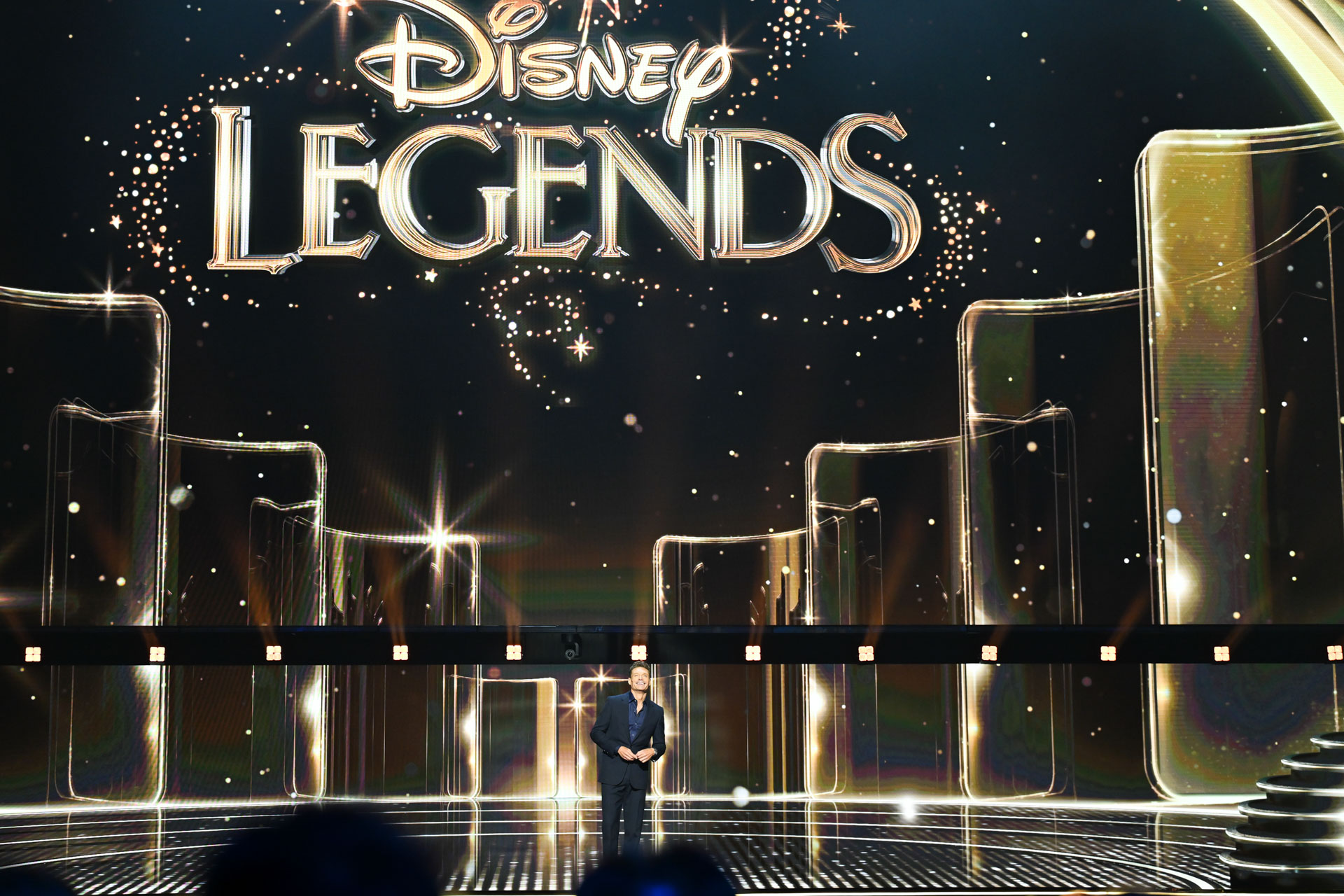 Ryan Seacrest stands onstage beneath a Disney Legends display while hosting the 2026 Disney Legends Awards ceremony.