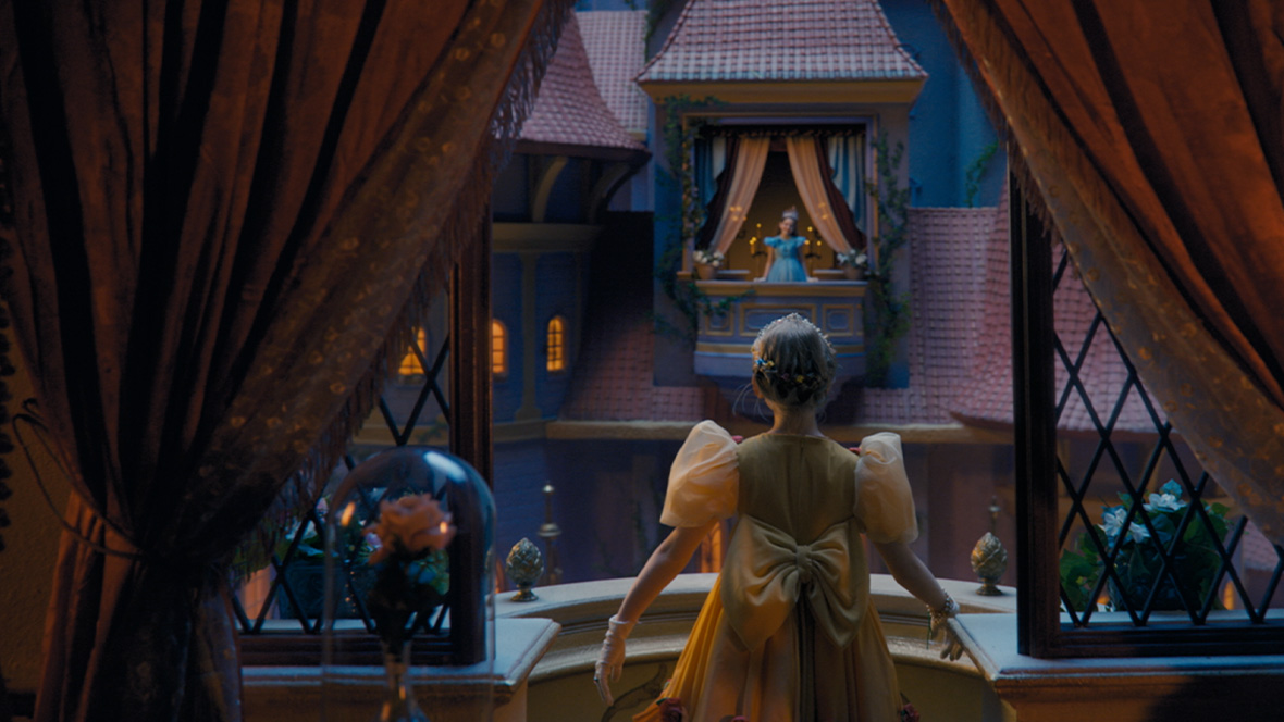 A person in a formal yellow gown with a large bow on the back stands at an open window framed by dark curtains, looking across a softly lit courtyard. In the distance, another figure stands on a decorated balcony of a castle-like building with peaked roofs and glowing windows. Warm indoor lighting contrasts with the cool blue tones of the evening scene, creating a fairy-tale atmosphere.