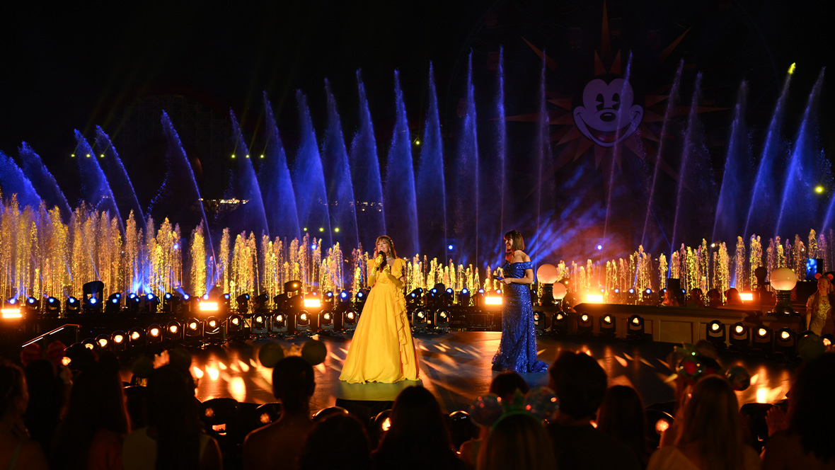 Two performers stand on an illuminated outdoor stage in front of a large audience. One performer wears a flowing yellow gown and the other a sparkling blue gown. Colorful fountains rise behind the stage, lit in blue, gold, and purple, while bright spotlights and decorative lighting create a dramatic nighttime backdrop. A large stylized character face is visible above the fountains, and audience members are silhouetted in the foreground.