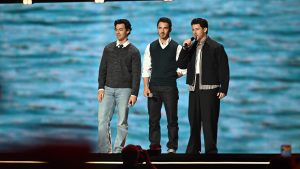 An image of Joe, Kevin, and Nick Jonas, on stage at the Honda Center during the Disney Entertainment Showcase at D23: The Ultimate Disney Fan Event, telling the crowd about Camp Rock 3.