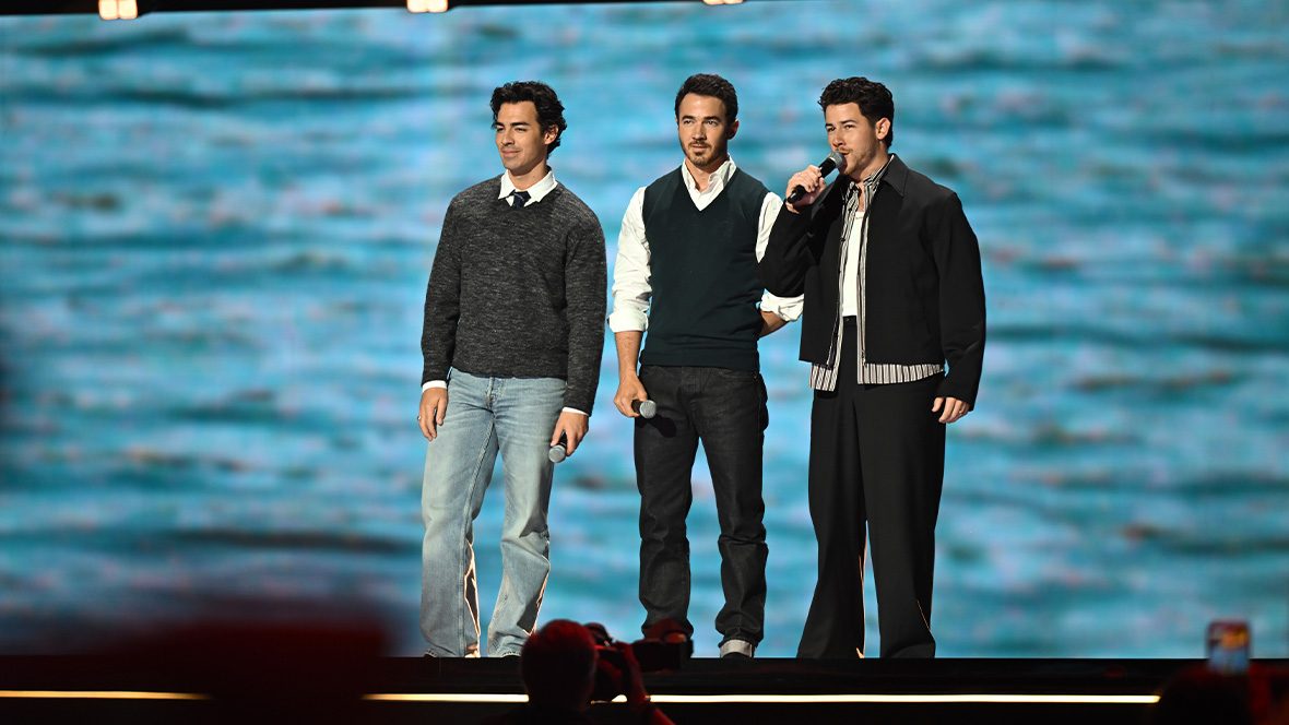 An image of Joe, Kevin, and Nick Jonas, on stage at the Honda Center during the Disney Entertainment Showcase at D23: The Ultimate Disney Fan Event, telling the crowd about Camp Rock 3.