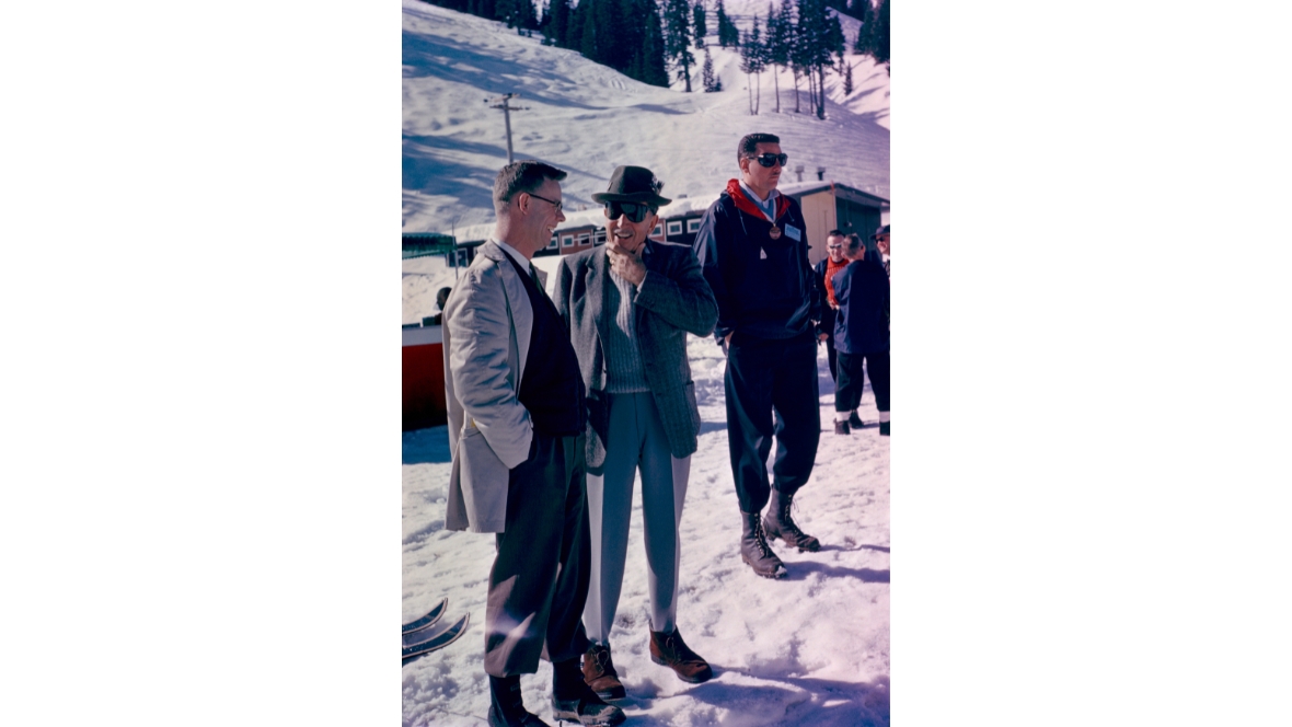 An image of Walt Disney visiting Sugar Bowl Ski Resort near Lake Tahoe. He is speaking to someone to his left, and other man are seen to his right. Snowy hills and trees are seen in the background.