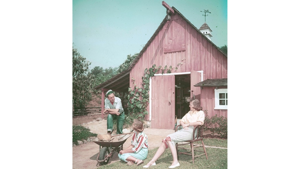 A color image of Walt Disney and his two daughters in their Holmby Hills backyard, with a reddish wooden barn seen in the background.