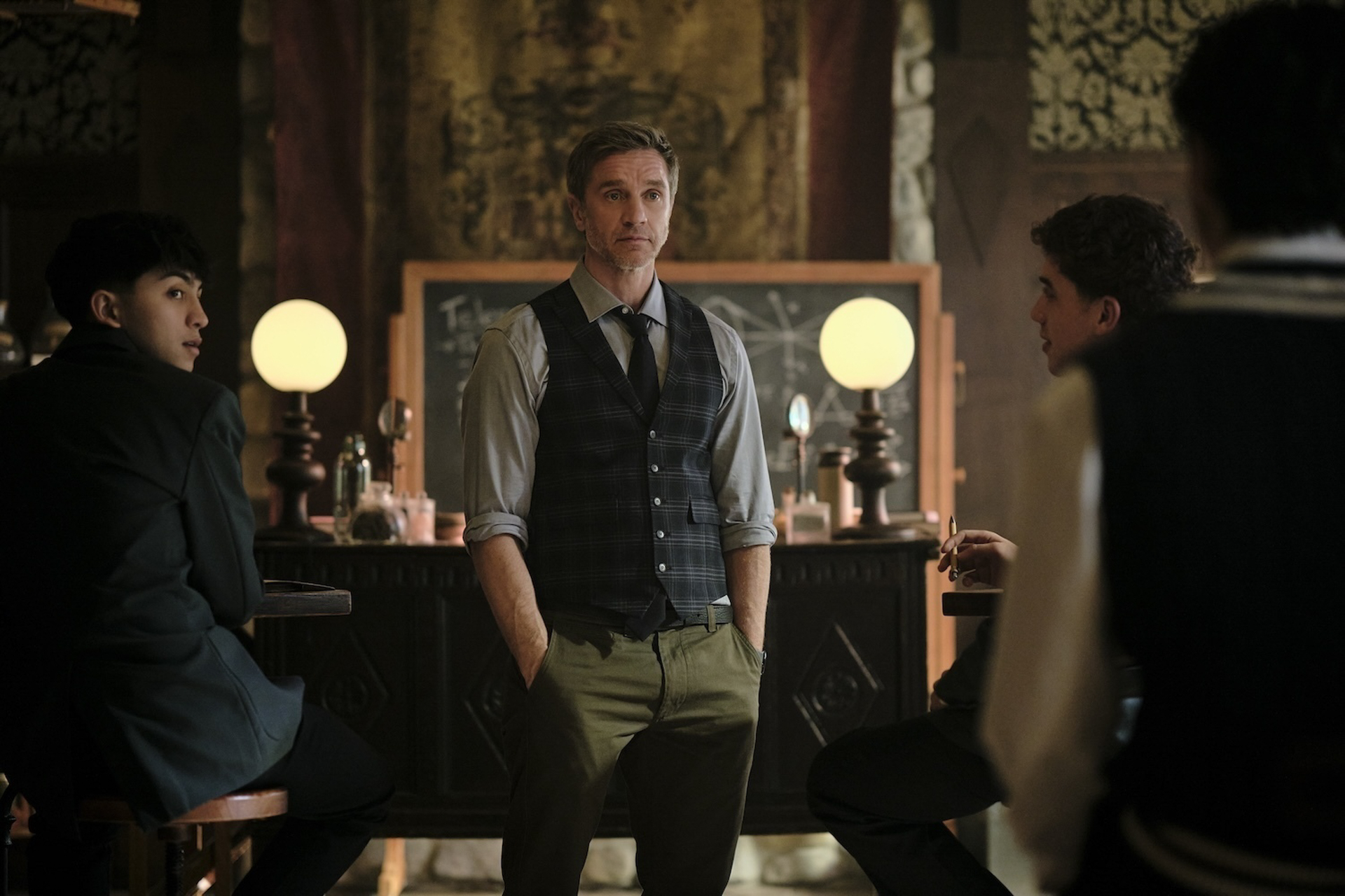 Mr. Cole, played by Devon Sawa, stands in the center of a classroom. Two students have their backs turned to him as they face someone out of frame in a scene from Coven Academy.