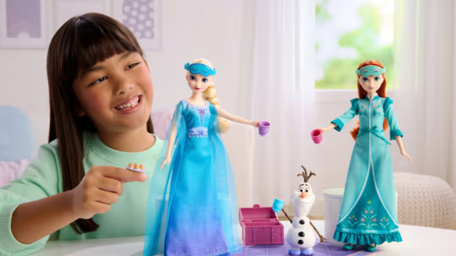 A child plays with Disney Frozen dolls at a table, holding a small toy dish beside dolls resembling Elsa, Anna, and Olaf with miniature play accessories.
