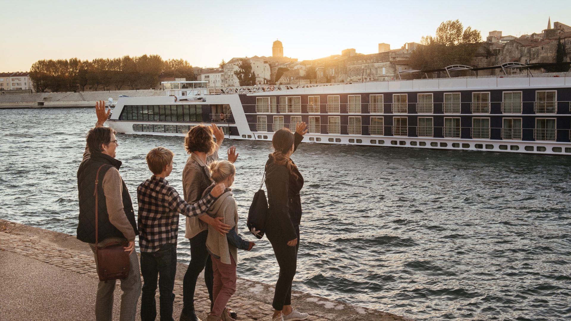 In a promotional image for Adventures by Disney, visitors wave toward a river cruise ship traveling along the Rh&ocirc;ne waterfront at sunset.