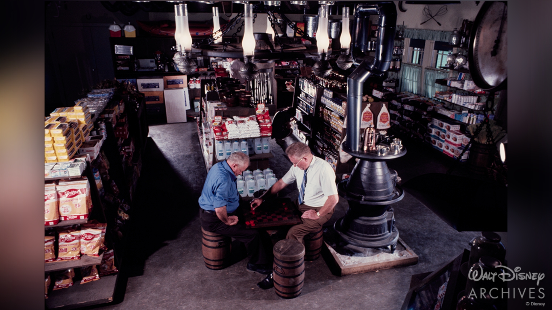 A rustic general store is stocked to the brim with a variety of provisions a family might need during their stay. Two men sit in the foreground of the image and play a game of checkers.